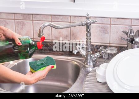 Frau mit roter Maniküre, die Waschmittel in die Geißel stellt, um die Gerichte zu waschen. Stockfoto