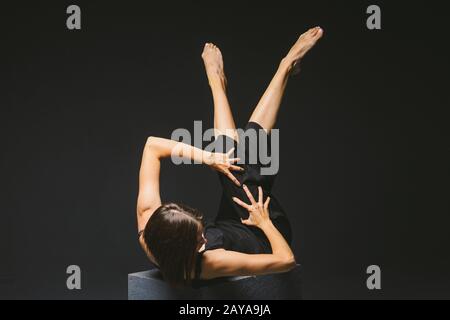 Gesellschaftliches Thema: Junge kaukasische Frau in seltsamer Pose im Kleid auf schwarzem Würfel, dunklem Hintergrund, symbolisiert Schmerz, Leid, Stockfoto