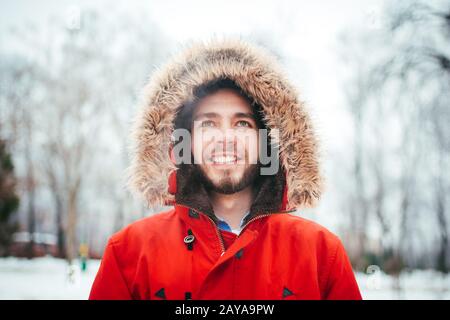 Porträt, Nahaufnahme eines jungen, stilvoll gekleideten Mannes, der mit einem Bart in einer roten Winterjacke mit Kapuze und Pelz auf h lächelt Stockfoto