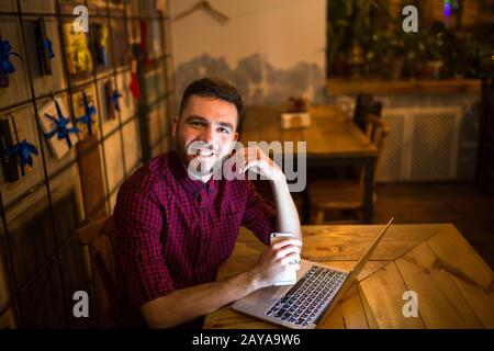 Ein junger, gutaussehender kaukasischer Mann mit Bart und tollem Lächeln in rotem Hemd arbeitet hinter dem Laptop, der auf der Holzlasche sitzt Stockfoto