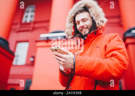 Gutaussehende junge männliche Studentin mit tollem Lächeln und Bart steht auf rotem Wandhintergrund, Fassade der Bildungseinrichtung in Rot Stockfoto