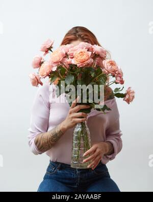 Eine Frau mit einer Tätowierung, die einen großen Blumenstrauß aus rosafarbenen Rosen in einer Glasvase um einen grauen Hintergrund mit Kopierraum hält. Mutter D Stockfoto