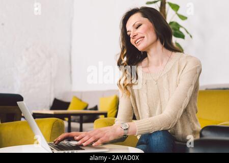 Junge kaukasische Frau mittleren Alters mit langen Haaren. Freizeitkleidung mit Laptop im Innenbereich. Café-Restaurant auf Holzstuhl Stockfoto