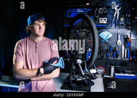 Thema ein kleines Unternehmen der Fahrradreparatur: Stilvoller Werkstattbesitzer steht in der Nähe der Maschine zur Reparatur des Rades auf der Rückseite Stockfoto