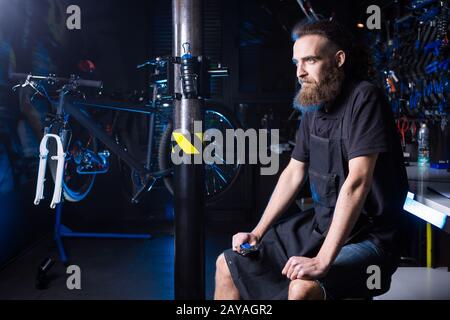 Porträt des Kleinunternehmers des jungen Mannes mit Bart. Kerl Fahrradmechaniker Werkstattarbeiter sitzt mit Werkzeug in der Hand in Stockfoto