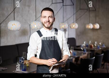 Ein junger, lächelnder Kellner in einem Restaurant, der neben den Tischen mit einem Glas Wein steht. In eine Schürze gekleidet, wird eine Id nehmen Stockfoto