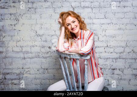 Nahaufnahme Porträt Schöne junge Studentin mit roten lockigen Haaren und frechem Gesicht auf einer Ziegelwand in grauer BA sitzend Stockfoto