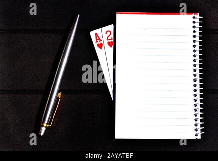 Stift mit Notizbuch und Spielkarten über klassischem Holztisch. Schreibgeräte, die oben auf dem schwarzen Hintergrund platziert sind. Artis Stockfoto