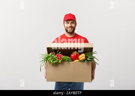 Lieferung Konzept: Stattlichen kaukasischen Lebensmittel-lieferservice Kurier Mann in der roten Uniform mit Lebensmittelgeschäft, mit frischem Obst und Gemüse Stockfoto