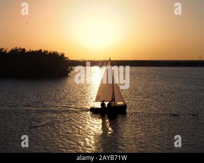 Ein Boot, das auf einem See auf dem Weg eines goldenen Sonnenuntergangs mit unidentifizierbaren Menschen in Silhouette segelt und im c schwimmt Stockfoto