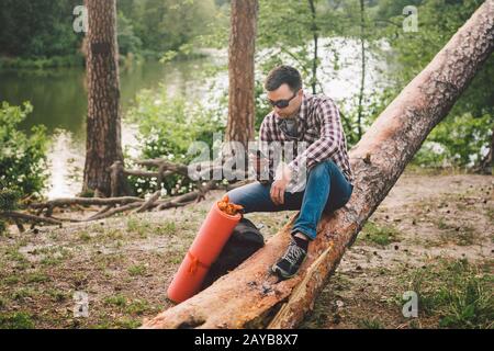 Der junge Mann sitzt auf dem Lastwagen im Wald und benutzt das Handy. Der Reisende sitzt auf einem großen umgestürzten Baum und hält das Telefon in der Hand. Thema Hi Stockfoto