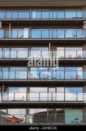 Die Reihen der Apartments in einem großen, modernen Gebäude mit gläsernen Balkons und Gartenmöbeln und Himmel spiegeln sich in den Fenstern wider Stockfoto