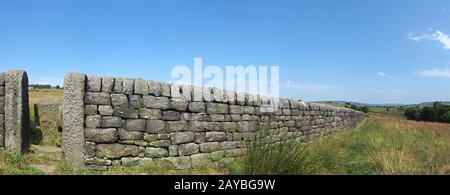 Lange Sicht auf eine Steinmauer mit Tor zwischen grasbedeckten Sommerfeldern mit blauem Himmel in der Landschaft von West yorkshire Stockfoto