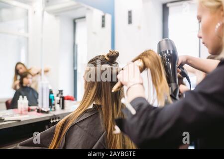 Porträt einer glücklichen Frau im Friseursalon. Professionelles Haar-Styling-Konzept. Friseursalon trocknet Mädchen lange Haare mit Haarfön Stockfoto