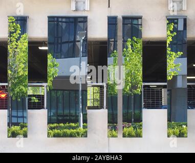 Eine städtische Straßenszene mit modernem Bürogebäude und Bäumen, die sich in gespiegelten Flächen auf einer Parkhausstruktur widerspiegelt Stockfoto