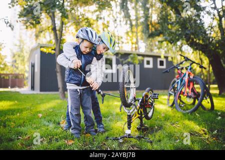 Zwei Kinder, ältere Jungen und jüngere Bruder lernen Reparaturrad. Zwei Jungs Geschwister in Helmen und Einzelkleidung benutzen Pumpwerkzeug Stockfoto