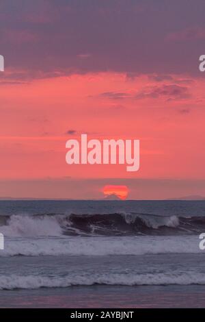 Sonnenuntergang in Jaco Beach, Costa Rica. Wellen im Vordergrund und die Sonne, die sich hinter einem kleinen Berg versteckt. Stockfoto