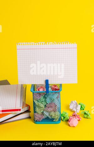 Kleiner Papierkorb voller zerknittertem farbigem Papier und einer blauen Wäscheklammer, die eine bunte Note in einem gelben Hintergrund aufhängen. Bürosup Stockfoto