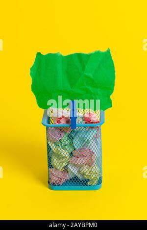 Kleiner Papierkorb voller zerknittertem farbigem Papier und einer blauen Wäscheklammer, die eine bunte Note in einem gelben Hintergrund aufhängen. Bürosup Stockfoto