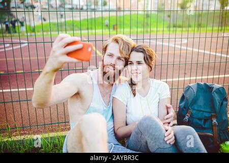 Ein Paar roter Haare und ein Bart eines europäischen Mannes und eine Frau ruhen, nachdem sie im Freien Sport gespielt haben. Der Kerl hält ein rotes Smartphone an Stockfoto