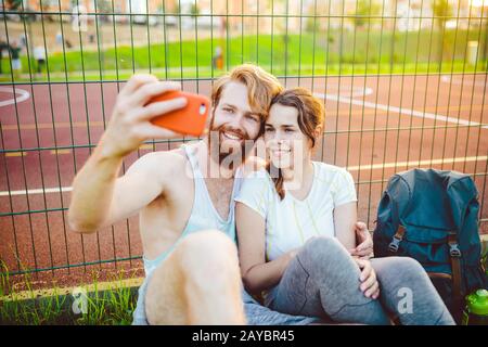 Ein Paar roter Haare und ein Bart eines europäischen Mannes und eine Frau ruhen, nachdem sie im Freien Sport gespielt haben. Der Kerl hält ein rotes Smartphone an Stockfoto