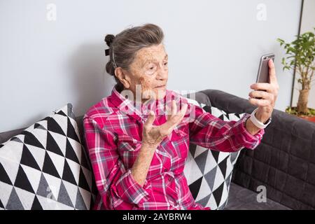 Eine sehr alte kaukasische Großmutter mit grauem Haar und tiefen Falten sitzt zu Hause auf der Couch in Jeans und rotem kariertem Hemd und Stockfoto