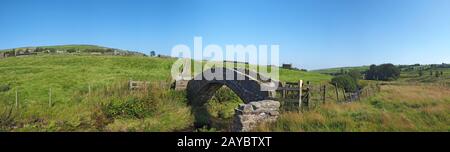 Ein Panoramablick auf eine alte Steinpackerbrücke, die einen Bach in der Landschaft West Yorkshire Dales mit dem Dorf überquert Stockfoto