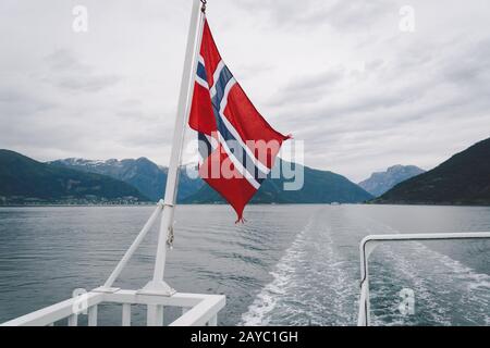 Norwegische Flagge, die am Geländer des Schiffes hängt und über dem Wasser winkt. Nordegischer Fjord mit einer Flagge. Fährfahrt in Norwegen. Stockfoto