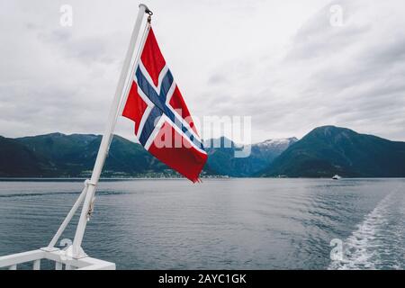 Norwegische Flagge, die am Geländer des Schiffes hängt und über dem Wasser winkt. Nordegischer Fjord mit einer Flagge. Fährfahrt in Norwegen. Stockfoto