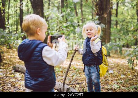 Kindervorschuler kaukasischer Bruder und Schwester machen im Herbst mit der Handy-Kamera im Waldpark Fotos voneinander Stockfoto