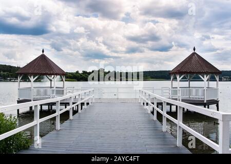 Weißer, überdachter Pier mit Holzhütten am See, grünem Wald und Wolken im Hintergrund, Polen Stockfoto