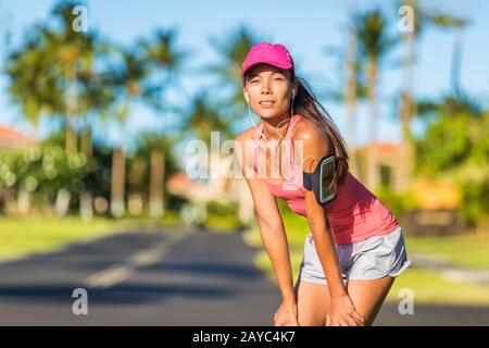 Müde Läuferin bereit zum laufen mit Sportkappe und Sportarmband mit Ohrhörern, die mobile Musik hören. Active Fit Asian Mädchen, die sich auf der Sommerstraße im Freien ausruhen und eine Joggingpause einlegen. Stockfoto