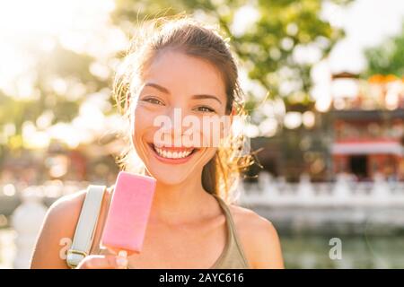 Fröhlicher Sommer verwöhnen Sie asiatisches Mädchen, das Erdbeer-Eis-Stick draußen bei Sonnenuntergang im Sonnenschein isst. Lächelnde Chinesin lächelt am Leben im Stadtpark. Stockfoto