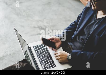 Der Geschäftsmann sitzt mit Smartphone auf dem Zementboden und verwendet einen Laptop Stockfoto
