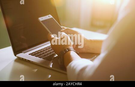 Silhouette eines zugeschnittenen Schusses eines jungen Mannes, der mit einem Smartphone und einem Notebook von zu Hause aus arbeitet Stockfoto