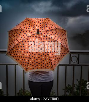 Teenager-Mädchen hält einen pfirsichfarbenen Regenschirm mit Punktmuster und genießt die Regenzeit von Monsoons inmitten dichter Wolken an einer Klippe Stockfoto