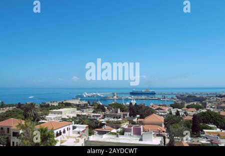 Ein Panoramablick auf die stadt rhodos mit Gebäuden der Stadt und alten Mauern rund um den Hafen mit Schiffen und Booten neben einem blauen s Stockfoto