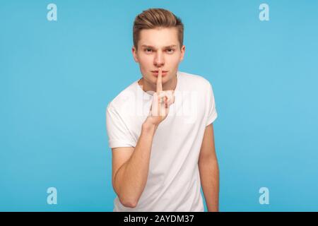 Shh, sei ruhig! Porträt eines ernsthaften Mannes in legeren weißen T-Shirts, das mit dem Finger auf den Lippen zur Kamera schürte, sich auflegte, nicht mehr spricht und Silenc hält Stockfoto