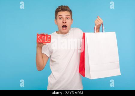 Überraschter freudiger Mann mit Verkaufsschrift und Einkaufstaschen, der mit Erstaunen vor der Kamera blickt, schockiert von günstigen Rabatten, schwarzer freitag. In Stockfoto