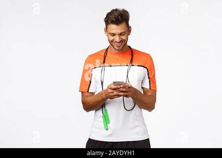 Sport-, Technologie- und Trainingskonzept. Gutaussehender junger Mann überprüft Fitness-App, hält Smartphone, lächelt wie beim Tippen am Gerät, wr Stockfoto