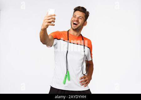 Gutaussehende, männliche Athletin nimmt selfie nach dem Training im Fitnessstudio, hält Smartphone mit verlängertem Arm, lächelt zufrieden und stolz, erzählt Stockfoto