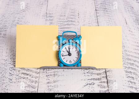 Mini-Blauer Farb-Wecker, der die Zeit zwischen zwei gelben Notizbüchern zeigt. Klassische kleine Uhr, die über einem Puffer steht Stockfoto