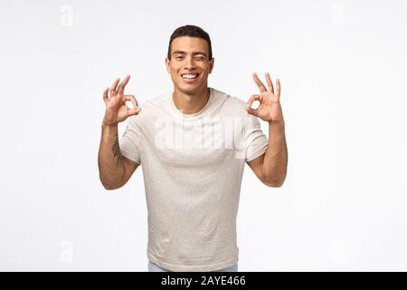 Frech sorglos und unbehelligt junger hispanischer Musklinenmann im T-Shirt, der in Ordnung zeigt, kein Problem oder perfekte Geste, Augenzwinkern und Schmieren Stockfoto