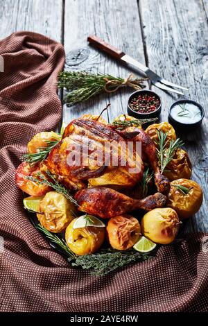 Gebratenes Hähnchen auf einer schwarzen Platte mit gebackenen Äpfeln und aromatischen Kräutern auf einem rustikalen Holztisch mit Gabel und Gewürzen, vertikaler Blick von Stockfoto
