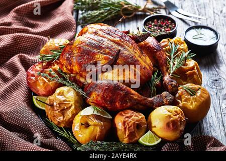Nahaufnahme eines gebratenen Hühnchens auf einer schwarzen Platte mit gebackenen Äpfeln und aromatischen Kräutern auf einem rustikalen Holztisch mit Gabel und Gewürzen, Hori Stockfoto