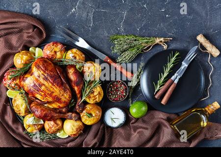 Nahaufnahme eines ganzen Rotisserie Huhns auf einer schwarzen Platte mit gebackenen Äpfeln und aromatischen Kräutern auf einem Betontisch mit Gabel und Gewürzen, Horizo Stockfoto
