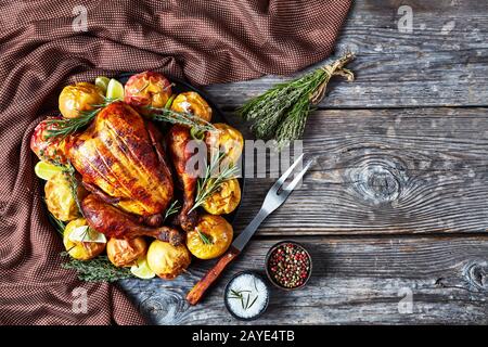 Gebratenes Hähnchen auf einer schwarzen Platte mit gebackenen Äpfeln und aromatischen Kräutern auf einem rustikalen Holztisch mit Gabel und Gewürzen, horizontaler Ansicht fr Stockfoto