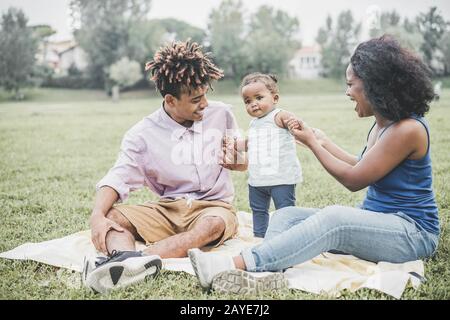 Fröhliche schwarze Familie, die Spaß hat, im Freien Picknick zu machen - Eltern und ihre Tochter genießen an einem Wochenendtag Zeit zusammen - Liebe zärtliche Momente und Happ Stockfoto