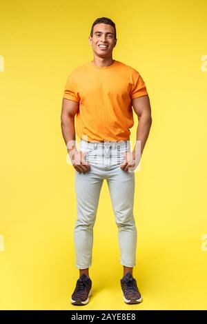 Vertikales, durchgehendes Studio schoss jungen Flusspferd mit Gefäßkörper, stehendem orangefarbenem T-Shirt und weißen Hosen, halten Sie die Hände in Po Stockfoto
