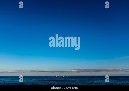 Segelboote am Horizont säumen das Meer, mit Blick auf den Strand von Barcelona (Spanien) an einem lauwarmen Winternachmittag. Verschiedene Blautöne. Stockfoto